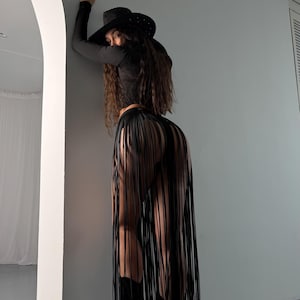 Puede incluir: Una mujer con una camisa negra de manga larga, un sombrero vaquero negro y una falda negra con flecos. Est&aacute; de pie frente a una pared blanca.