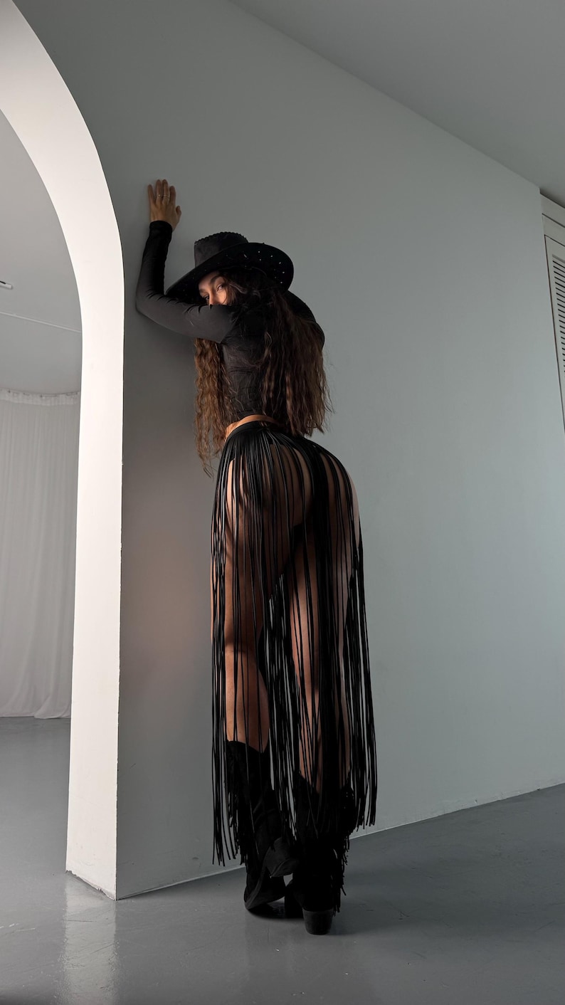 Puede incluir: Una mujer con una camisa negra de manga larga, un sombrero vaquero negro y una falda negra con flecos. Est&aacute; de pie frente a una pared blanca.