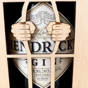Könnte beinhalten: Ein Holzrahmen mit einer Flasche Hendrick's Gin darin. Der Rahmen hat ein gefängnisähnliches Design mit zwei Fäusten, die die Stäbe halten. Die Fäuste haben die Wörter "TOUGH" und "LIFE" tätowiert. Das Etikett der Gin-Flasche ist sichtbar.