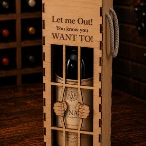 Könnte beinhalten: Eine Weinflaschenbox aus hellem Holz im Gefängniszellen-Design mit vertikalen Stäben und zwei hölzernen Händen, die diese umklammern. Der Text Let me Out! You know you WANT TO! ist vorne eingraviert. Ein grauer Seilgriff ist seitlich angebracht.