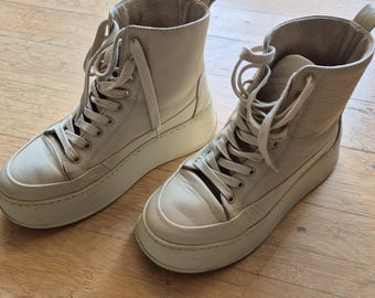 Puede incluir: Un par de zapatillas altas de color beige claro. Las zapatillas tienen suelas gruesas, cordones y un cuello acolchado. Los zapatos están hechos de un material liso y parecen nuevos. Las zapatillas están sobre una superficie de madera.