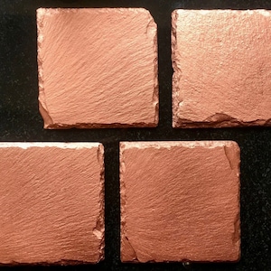 May include: Four square copper-coloured slate coasters with rough, textured edges. The coasters have a metallic sheen and a natural, rustic appearance. Ideal for protecting surfaces from drink condensation.