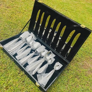 Könnte beinhalten: Ein Besteckset aus silberfarbenem Edelstahl in einem schwarzen Aufbewahrungskoffer. Das Set enthält acht Messer in Schlitzen an der Rückseite sowie verschiedene Gabeln, Löffel und kleinere Utensilien, die in durchsichtige Kunststofffolie eingewickelt sind.