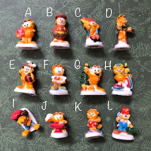 Peut inclure: Une collection de douze figurines Garfield miniatures, chacune représentant le chat de dessin animé dans différentes poses. Les figurines sont principalement orange, avec du blanc et d'autres couleurs d'accentuation. Chaque figurine est sur une petite base blanche. Le fond est vert.