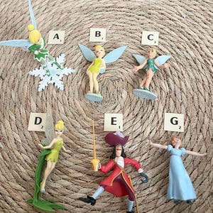May include: A collection of miniature figurines featuring characters from Peter Pan. The figurines include Tinkerbell in various poses, Captain Hook, and Wendy. The figures are brightly colored and set against a woven background.