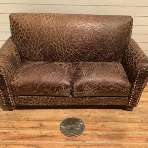 May include: A brown leather sofa with tufted upholstery and gold-colored nailhead trim. The sofa has a distressed look and is sitting on a wooden floor.