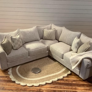 Peut inclure: Un canapé d'angle gris clair avec plusieurs coussins et une couverture crème. Le canapé est posé sur un sol en bois avec un tapis rond beige à bord festonné. La pièce a une palette de couleurs neutres.