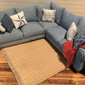 May include: A blue denim sectional sofa with a variety of throw pillows, including a white pillow with a navy blue star, a white pillow with French text, and a blue pillow with a geometric pattern. A red throw blanket is draped over the arm of the sofa. A tan rug with fringe is in front of the sofa.
