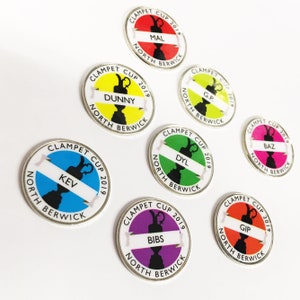 May include: Eight round, silver-coloured pins with colourful backgrounds and black silhouettes of a trophy cup. Each pin has the text "CLAMPET CUP 2019" and "NORTH BERWICK" around the edge. The names "MAL", "DUNNY", "KEV", "G.P.", "DYL", "BIBS", "BAZ", and "GIP" are printed on the pins.