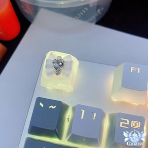 May include: A white keycap with a white bow and a clear rhinestone in the center. The keycap is glowing in the dark.