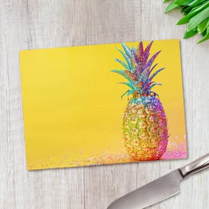 May include: A rectangular glass chopping board featuring a vibrant yellow background and a central pineapple graphic. The pineapple displays a multi-coloured, iridescent, and glittery finish with shades of blue, purple, pink, and gold throughout.