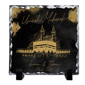 Puede incluir: Una placa cuadrada de pizarra negra con bordes plateados irregulares, que presenta una ilustraci&oacute;n dorada de la Kaaba. El texto Umrah Mubarak, Imran & Zeena y March 2023 aparece en letra dorada. La placa se sostiene sobre dos soportes negros.
