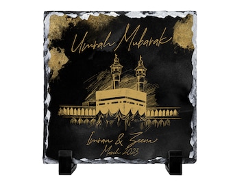 May include: A square black slate plaque with rough, silver-painted edges, featuring a gold-colored illustration of the Kaaba. The text reads Umrah Mubarak, Imran & Zeena, and March 2023 in gold script. The plaque is supported by two small black stands.