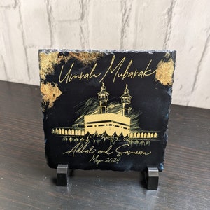 Puede incluir: Una placa cuadrada de pizarra negra con texto en color dorado que dice Umrah Mubarak, Adhal and Sameera, y May 2024. El centro muestra una ilustraci&oacute;n dorada de la Kaaba con dos minaretes, acentuada con detalles dorados en los bordes.