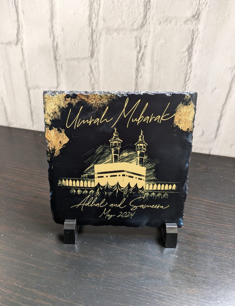Puede incluir: Una placa cuadrada de pizarra negra con texto en color dorado que dice Umrah Mubarak, Adhal and Sameera, y May 2024. El centro muestra una ilustraci&oacute;n dorada de la Kaaba con dos minaretes, acentuada con detalles dorados en los bordes.