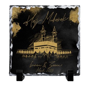 Puede incluir: Una placa cuadrada de pizarra negra con bordes irregulares pintados en plata. Presenta un boceto dorado de la Kaaba, el texto "Hajj Mubarak", los nombres "Imran & Zeena" y la fecha "March 2023" en letra dorada, sobre dos soportes negros.