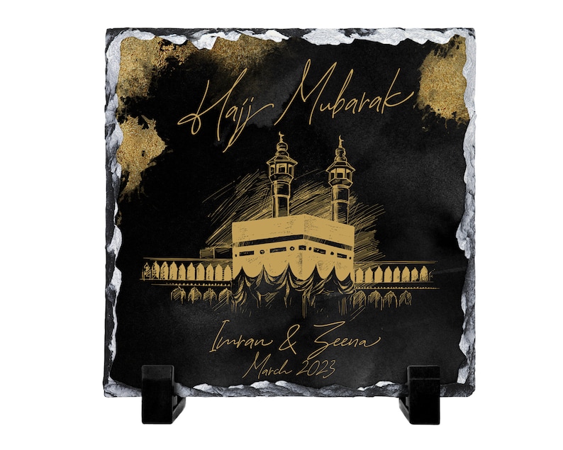 Puede incluir: Una placa cuadrada de pizarra negra con bordes irregulares pintados en plata. Presenta un boceto dorado de la Kaaba, el texto "Hajj Mubarak", los nombres "Imran & Zeena" y la fecha "March 2023" en letra dorada, sobre dos soportes negros.