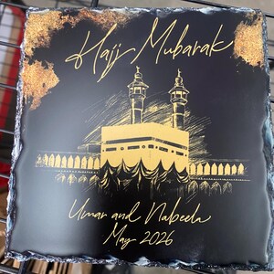 Puede incluir: Un posavasos cuadrado de pizarra negra con bordes irregulares. La superficie presenta una ilustraci&oacute;n dorada de la Kaaba con dos minaretes. El texto dice Hajj Mubarak, Umar and Nabeela, y May 2026 en una fuente dorada.