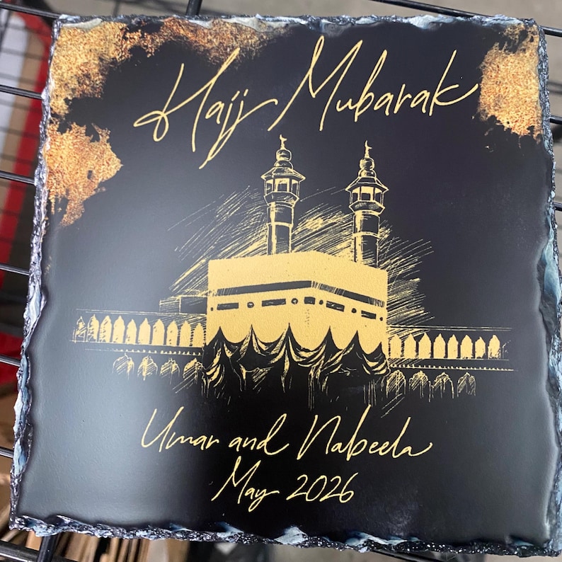 Puede incluir: Un posavasos cuadrado de pizarra negra con bordes irregulares. La superficie presenta una ilustraci&oacute;n dorada de la Kaaba con dos minaretes. El texto dice Hajj Mubarak, Umar and Nabeela, y May 2026 en una fuente dorada.
