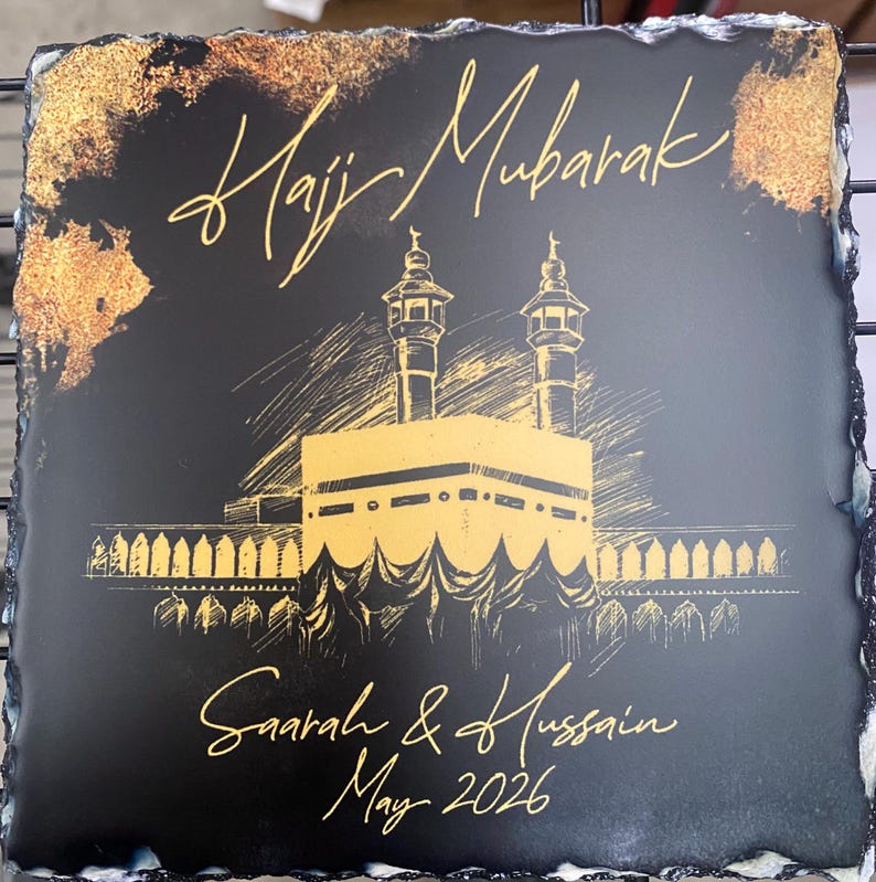 Puede incluir: Una placa cuadrada de pizarra negra con bordes irregulares que presenta ilustraciones y texto en tinta dorada. El dise&ntilde;o incluye el texto Hajj Mubarak, un boceto de la Kaaba con dos minaretes, y los nombres Saarah & Hussain junto a May 2026.