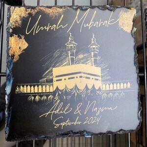 Puede incluir: Un posavasos cuadrado de pizarra negra con bordes irregulares. Presenta un dibujo lineal de color dorado de la Kaaba y minaretes. El texto dice Umrah Mubarak, Adil & Nazma y September 2024 en letra dorada, con detalles en pan de oro.