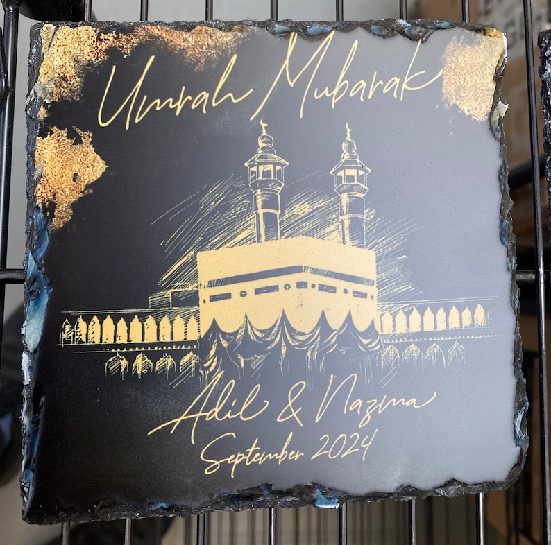 Puede incluir: Un posavasos cuadrado de pizarra negra con bordes irregulares. Presenta un dibujo lineal de color dorado de la Kaaba y minaretes. El texto dice Umrah Mubarak, Adil & Nazma y September 2024 en letra dorada, con detalles en pan de oro.