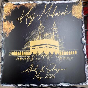 Puede incluir: Una placa cuadrada de pizarra negra con bordes naturales y rugosos. Presenta ilustraciones en tinta dorada de la Kaaba y dos minaretes. El texto dice Hajj Mubarak, Abid & Shazia y May 2026 en una fuente cursiva dorada con detalles de pan de oro.