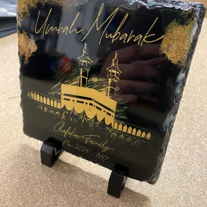 Puede incluir: Una placa cuadrada de pizarra negra con bordes naturales rugosos y acabado brillante. El dise&ntilde;o incluye el texto dorado Umrah Mubarak, Chafekar Family, Nov 2025 - 1447, junto con una ilustraci&oacute;n dorada de la Kaaba y los minaretes.