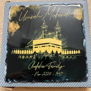 Puede incluir: Una placa cuadrada de pizarra negra con acabado brillante que presenta una ilustraci&oacute;n dorada de la Kaaba y minaretes. El texto dice Umrah Mubarak, Chafekar Family y Nov 2025 - 1447 en letra dorada. La placa tiene bordes naturales y rugosos.