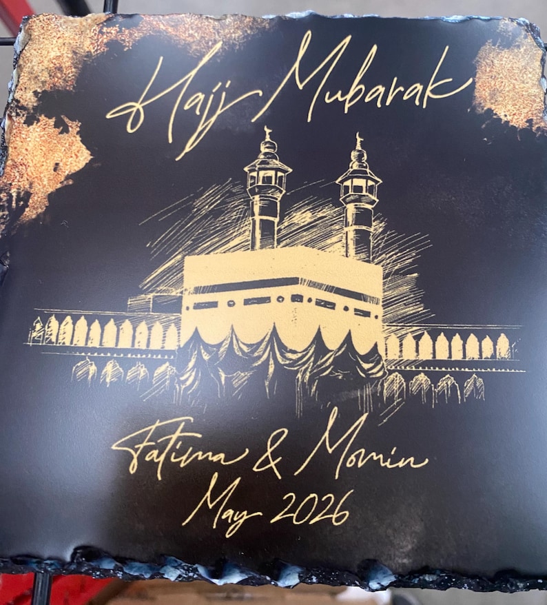 Puede incluir: Un posavasos cuadrado de pizarra negra con un boceto dorado de la Kaaba y dos minaretes. El texto "Hajj Mubarak" est&aacute; impreso en la parte superior, con "Fatima & Momin" y "May 2026" escritos en letra dorada en la parte inferior.