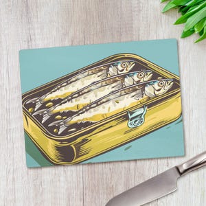 May include: Rectangular chopping board with a retro illustration of sardines in a tin. The sardines are shown with detailed scales and eyes, inside a yellow tin against a blue background. The board has a light blue border.