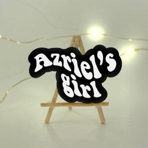 Może przedstawiać: Czarno-biała naklejka z tekstem "Azriel's girl" w retro czcionce. Naklejka jest wyświetlana na małym drewnianym sztalugach.