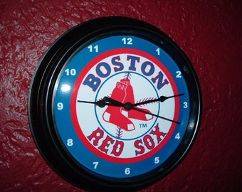May include: Round wall clock featuring the Boston Red Sox logo. The clock face has a blue outer ring with white numerals, a red and white center with the team logo, and black hands. The clock is in a black frame.