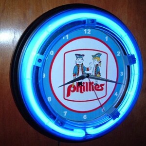 May include: Round neon clock with a blue neon light ring. The clock face features the Phillies logo with two cartoon figures and the word "Phillies" in red. The clock has black hands and a black border.