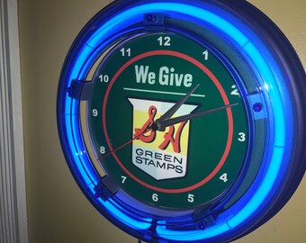 May include: A circular clock with a blue neon light surround. The clock face is green, displaying the words "We Give" and "GREEN STAMPS". Black clock hands indicate the time.