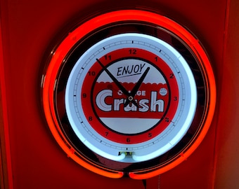May include: Orange Crush neon clock with a white face and black hands. The clock face features the words "Enjoy Orange Crush" in a red and white design. The clock is surrounded by a bright orange neon light.