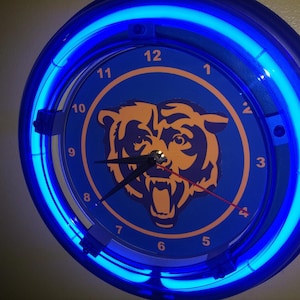 May include: A round wall clock with a blue face and a neon blue light ring. The clock face features a gold bear logo and black clock hands. The numbers are white. The clock is a sports-themed item.
