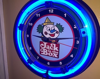 May include: A circular wall clock with a blue neon light surround. The clock face displays the Jack in the Box mascot, a smiling clown. The clock hands are black, and the second hand is red. The clock has the words "Jack in the Box" on the face.