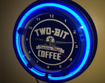 May include: A circular, dark blue neon clock with a bright blue neon border. The clock face displays the words "TWO-BIT" and "COFFEE" in white, with "Peaberry Blend" in a banner. The clock hands are black and red.