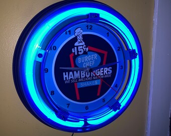 May include: A circular wall clock with a blue neon light surround. The clock face displays the text "Burger Chef," "Hamburgers," and "Shakes." The clock hands indicate approximately 3 o'clock. The clock is mounted on a light yellow wall.