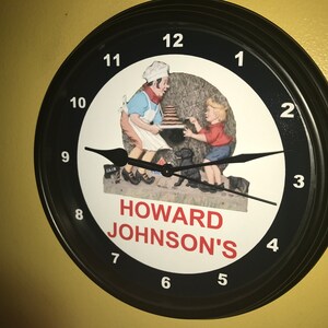 May include: Black-framed wall clock with a white face featuring a vintage illustration of a chef and a child. The clock hands are black, and the numbers are white. The text "HOWARD JOHNSON'S" is in red.