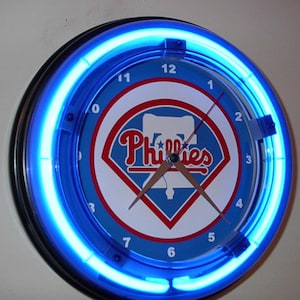 May include: A round wall clock with a blue neon light ring. The clock face features the Philadelphia Phillies logo in red, white, and blue. The clock hands are brown.