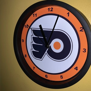 Peut inclure: Horloge murale ronde avec le logo des Flyers de Philadelphie en orange, blanc et bleu marine. L'horloge a un cadre noir et des chiffres orange. Les aiguilles sont noires, et l'heure est d'environ 11h00.