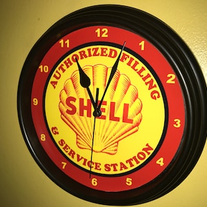 May include: A round wall clock with a black frame and a yellow and red face. The clock face features the Shell logo, a seashell, and the words "AUTHORIZED FILLING & SERVICE STATION". The clock hands are black.