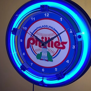 May include: A round, blue neon clock featuring the Philadelphia Phillies logo. The clock face displays the team's name in red script over a baseball graphic. The clock has a blue neon border and black clock hands.