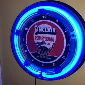 May include: A vintage Sinclair Pennsylvania Motor Oil clock with a blue neon light. The clock face is black with white numerals and a red and white Sinclair logo featuring a dinosaur. The clock is a circular shape.