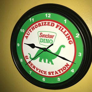 Op de afbeelding: Ronde wandklok met een zwart frame. De wijzerplaat is groen en wit en toont het Sinclair Dino-logo en de woorden "AUTHORIZED FILLING & SERVICE STATION". Zwarte wijzers geven de tijd aan.