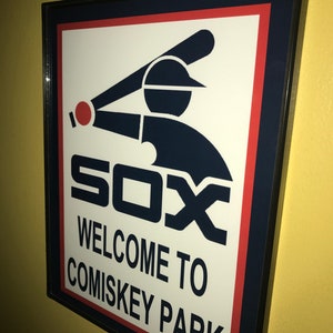 May include: Framed sign featuring the Chicago White Sox logo with a batter swinging a bat. The sign reads "SOX" and "WELCOME TO COMISKEY PARK" in black text on a white background, framed with navy blue and red.