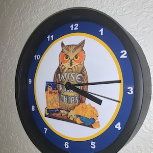 Könnte beinhalten: Runde schwarze Wanduhr mit blau-gelbem Zifferblatt. Die Uhr zeigt ein Vintage-Design von Wise Potato Chips mit einer Eule und einer Tüte Chips. Die Zeiger der Uhr sind schwarz.
