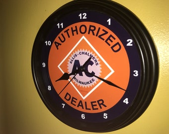 Puede incluir: Un reloj de pared redondo con marco negro. La esfera es naranja y azul marino, con el texto "AUTHORIZED DEALER" y "ALLIS-CHALMERS MILWAUKEE". Las manecillas del reloj son negras.